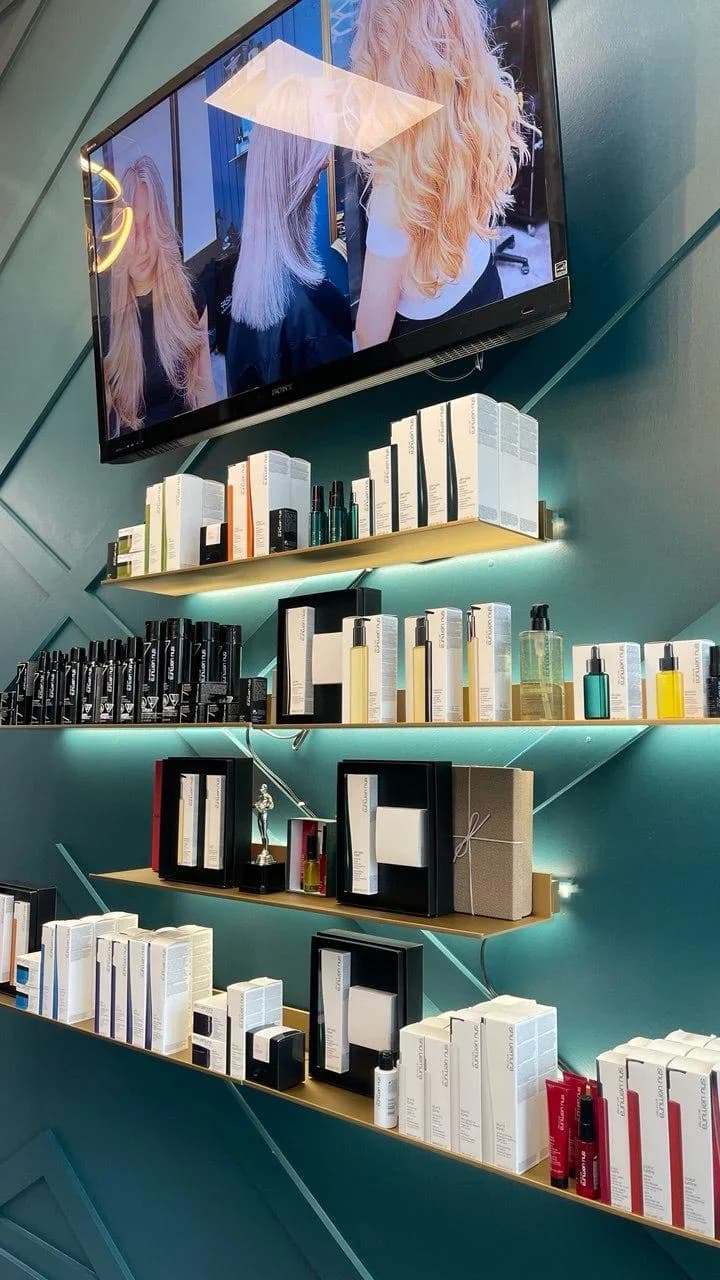 Backlit retail shelving on a teal wall holding haircare and skincare product ranges, with a screen above showing styling work