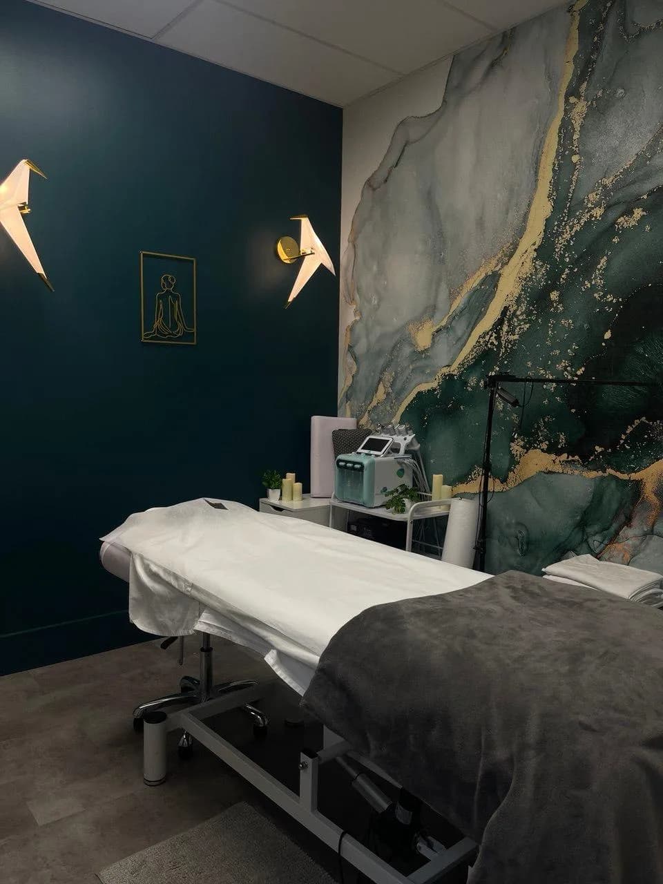 A private treatment room with a made-up facial bed, teal and gold marbled wall mural, and a facial treatment machine beside it